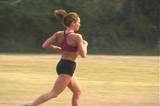 A beautiful young woman jogs in a grassy field.  Camera pans right to follow.
