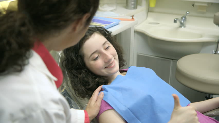 Camera pans down to cute girl in dentist chair getting approval from her dentist before smiling and displaying a thumbs up to someone off camera.