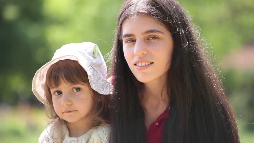 Brunette young adult mother talks to her little baby girl. Sunny day, outdoor portrait.
