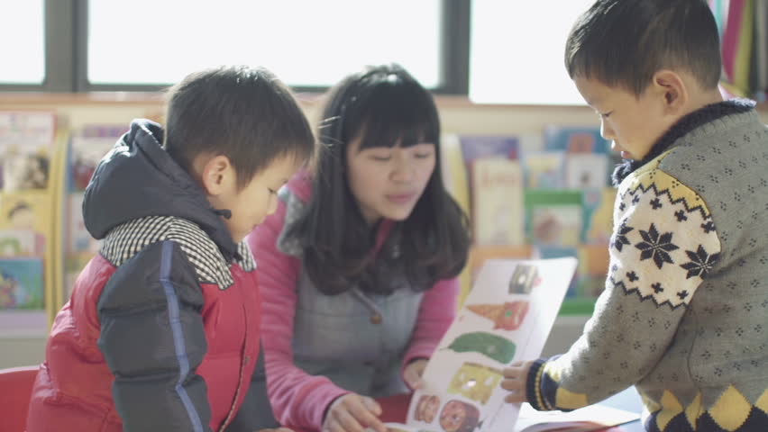 asian kindergarten teacher and her students sit a small table reading a book together in a classroom in china. 