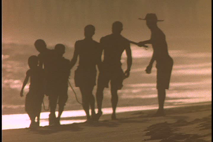 Slavery reenactment in Ghana, Africa. MS silhouette of overseer leading five men in chains (two are boys) up beach in very bright sunlight. 