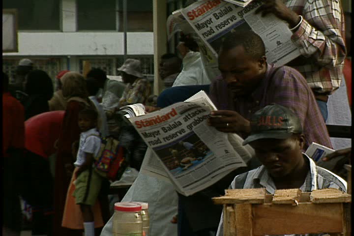 MOMBASA, KENYA, AFRICA - DECEMBER 01, 1998: MS Men reading newspapers on busy corner in crowded outdoor market. Many pedestrians walk past them.
