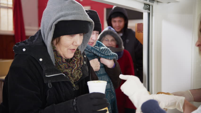 Friendly voluntary workers standing at a soup kitchen serving hatch are handing out cups of hot soup and bread to a waiting line of homeless and needy people. In slow motion