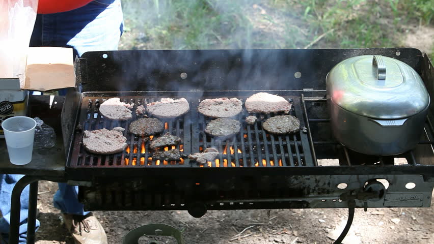 Close up of an outdoor grill cooking hamburgers during a BBQ. Hot fire and smoke. Large covered pot on warmer. Gas grill.