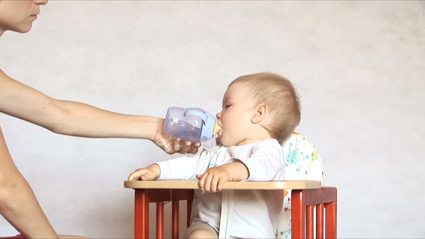 Baby sits at a children's little table. Mum gives to drink water of the child. Isolated on the white.