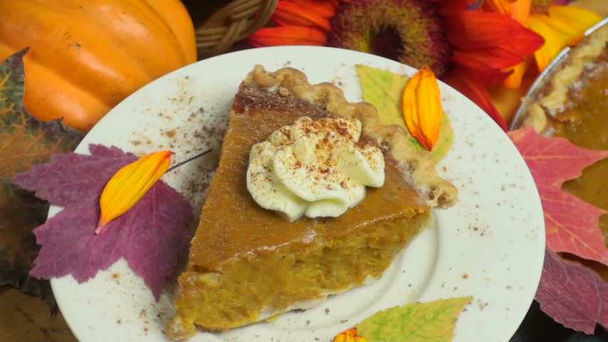 Slice of pumpkin pie, topped with whipped cream, displayed, rotating, above festive holiday background of whole pie, fall leaves, red sunflower, squash. 