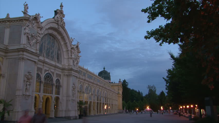 Pseudo-Baroque cast iron unique City Colonnade in Marianske Lazne (West Bohemia, Czech) with walking people and fountain during dusk and starting illumination. Time lapse.