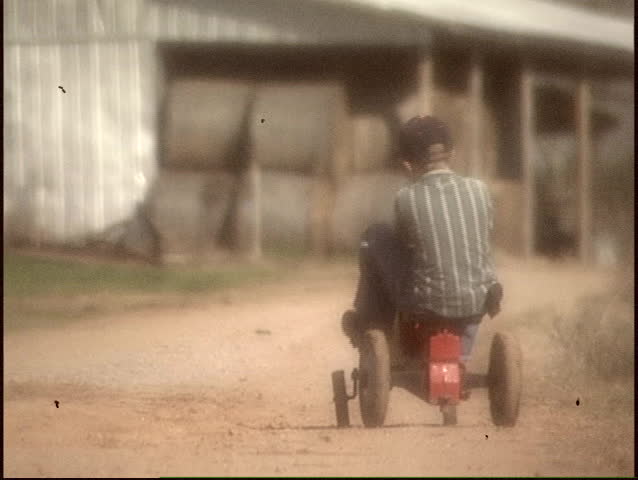 Slow motion - Vintage film look of a Rural farm boy (approximately 8 yrs. old) with a tractor pedal toy heading to the barn.