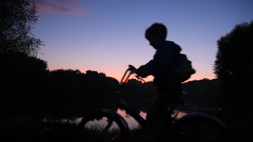 boy with rucksack on bicycle looks at sunset lake 