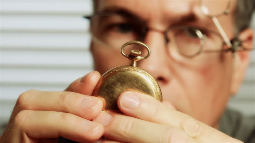 A man wearing jewelers glasses, picks up an old pocket watch and begins to examine it. Rack focus