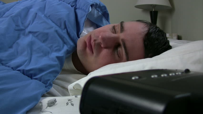 Man sleeping in bed and hits snooze, then gets up and tries again, gets mad and shoves alarm clock away.