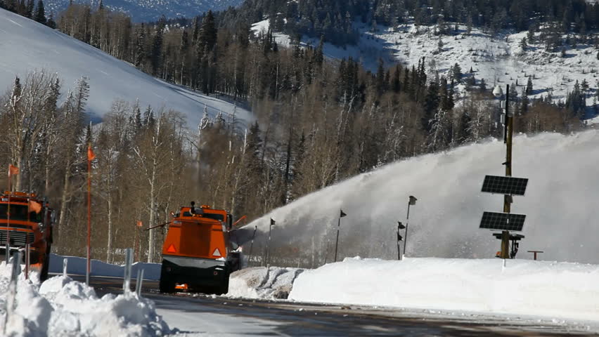 Snow blower and snowplow removing snow from mountain road. Large vehicle with spinning blades throws ice and snow off the road. 