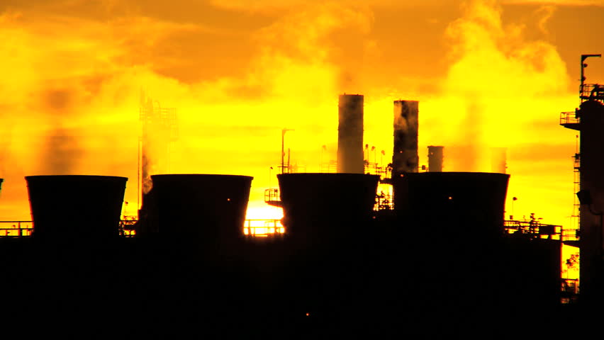 Smoke from oil refinery chimneys processing fossil fuel against setting sun