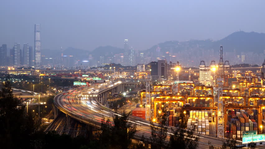 Busy City. Sunset Timescape. Hong Kong. Zoom out shot. Corporate buildings at the back. Busy cargo container loading activity in container terminal. Busy traffic across the main highway at rush hour.