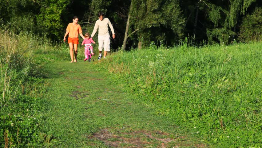 happy parents helps a little daughter riding bicycle to camera in summer park 