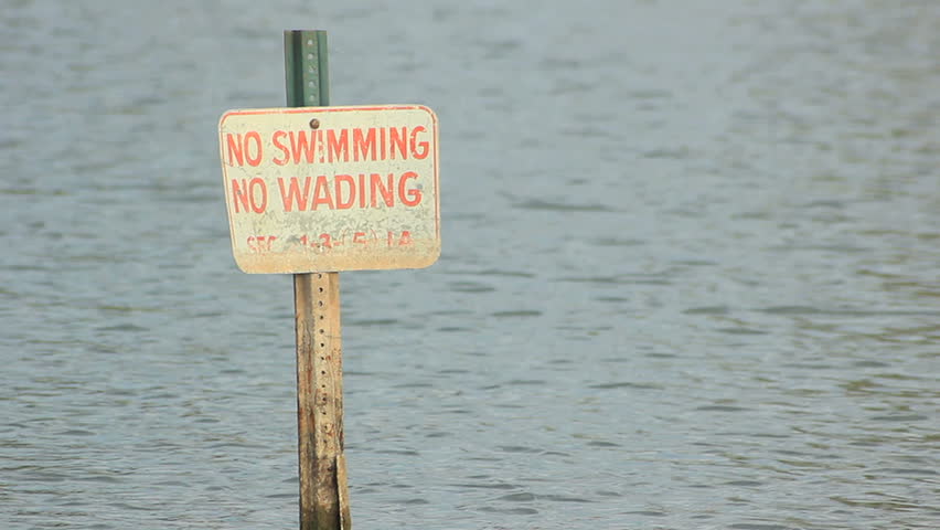 A warning sign reads "No swimming...no wading" while the waters move gently past the background. Long shot.