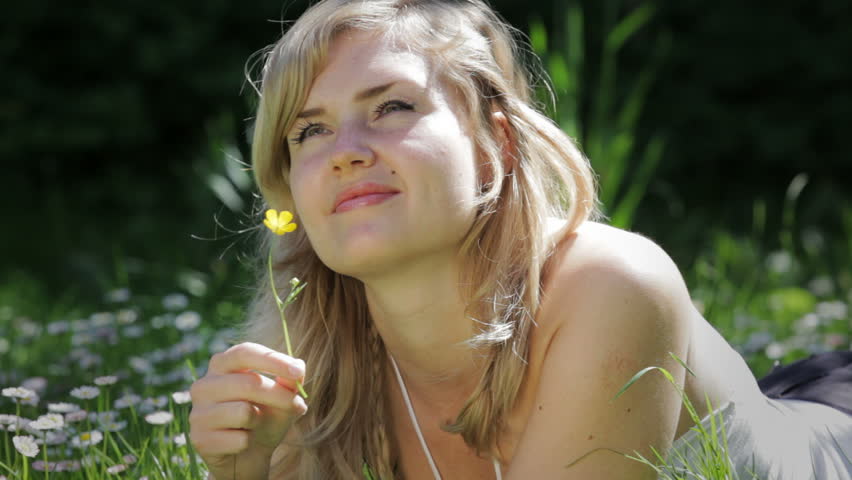 A young blond woman laying on the grass and smelling a flower
