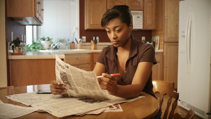 An African American woman performs various tasks around the house. She searches for a job, pays bills, makes coffee, studies the bible, reads a book, and works on her laptop.
