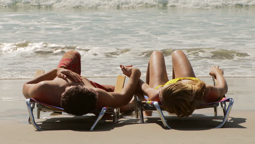 Young couple on lounge chairs holding hands by the ocean