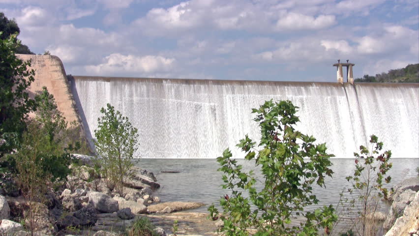 Video of a large diversion dam concrete spillway. Medina River in the Hill Country of south Texas. Tree lined hillside. Water flowing over top of dam. 