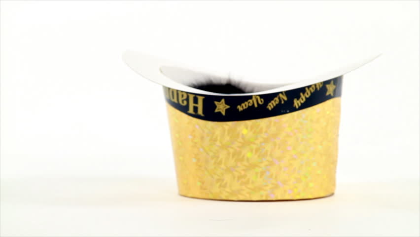 A tiny kitten inside of a gold colored Happy New Year fedora pokes his head up and then gingerly climbs out tipping the hat over. White backdrop
