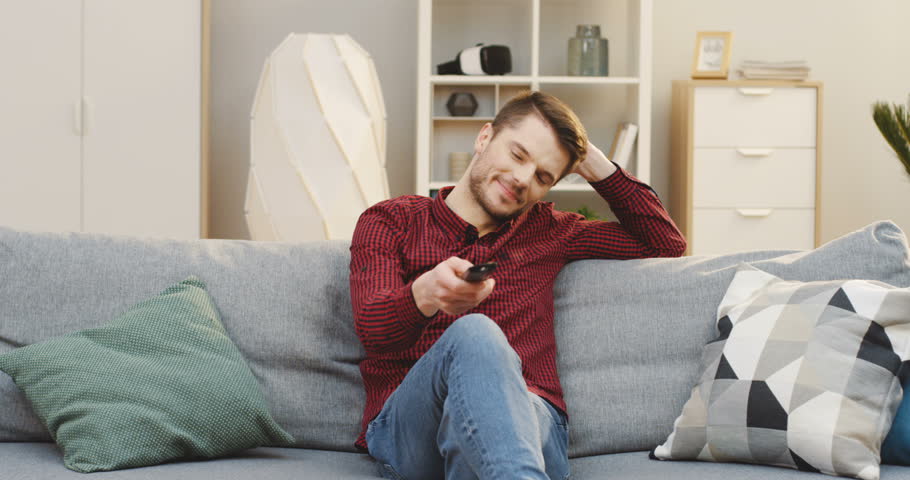 Young Handsome Man Sitting On The Sofa Watching Tv And Changing Channels In The Cozy Living Room At Home Indoor Young Handsome Man Sitting On The Sofa Watching Tv And Changing Channels In The Cozy Living Room At Home Indoor