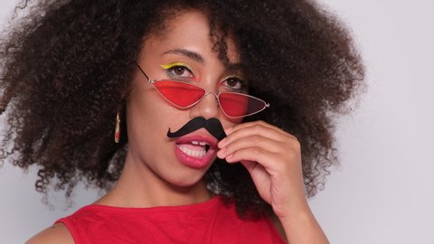 A Dark Skinned Model With Lush Black Hair In Red Glasses And A Red T Shirt Against A White Wall The Model Jokes With A Cardboard Mustache Showing His Masculine Character 4k Uhd Video