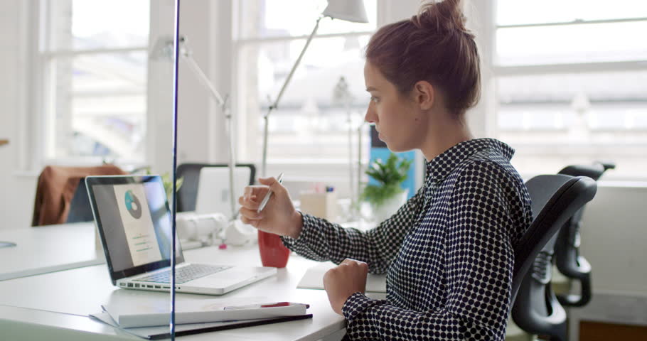 Young business woman working at shared desk in trendy hipster start up office using big data on laptop computer